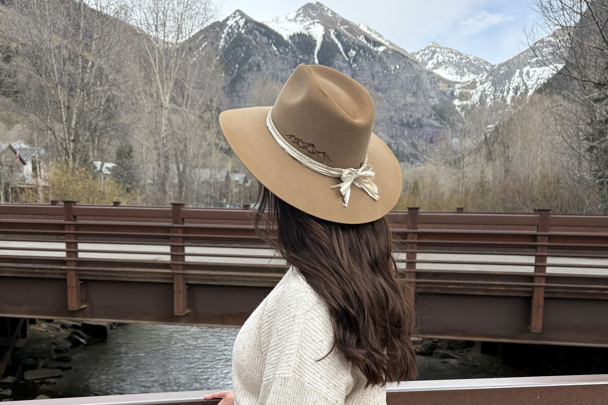 A woman in a camel felt hat with a cream ribbon band and a burned mountain-range detail, looking out at snowy peaks